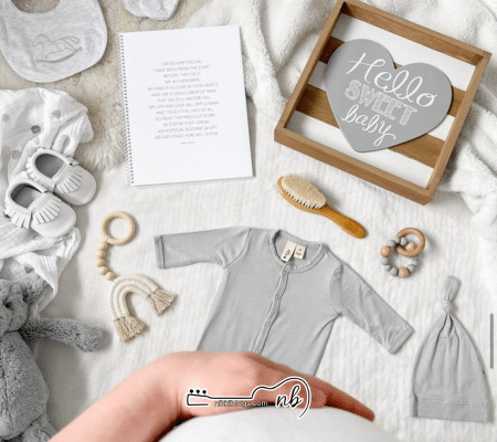 pregnant woman's hand holding her belly with a top down view of baby onesie, hat, shoes, brush, teether, swaddle, and other baby accessories