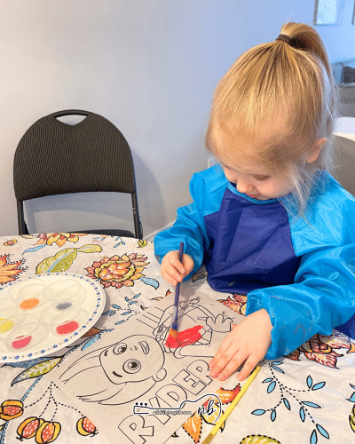 toddler painting with paintbrush using washable paints over table covered with outdoor table cloth